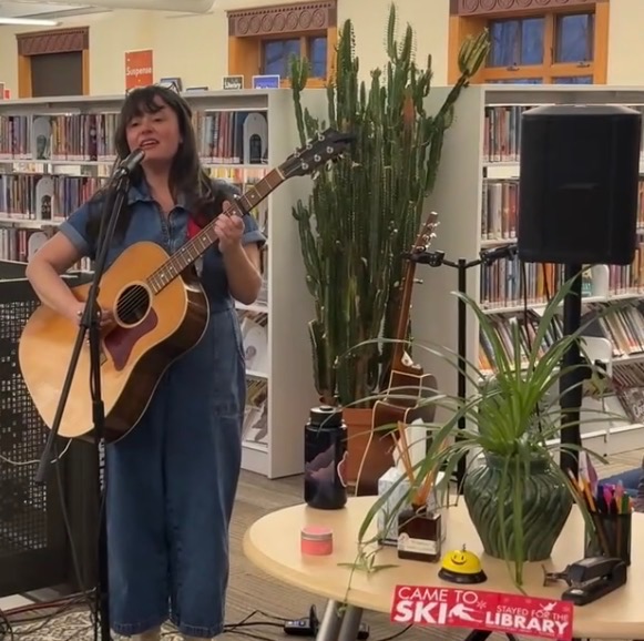 A woman plays guitar and sings into a microphone at the Wilkinson Public Library, with a large cactus nearby, during one of the Tiny Circ Desk Concerts. A table displays library materials and a Came to Ski sticker.
