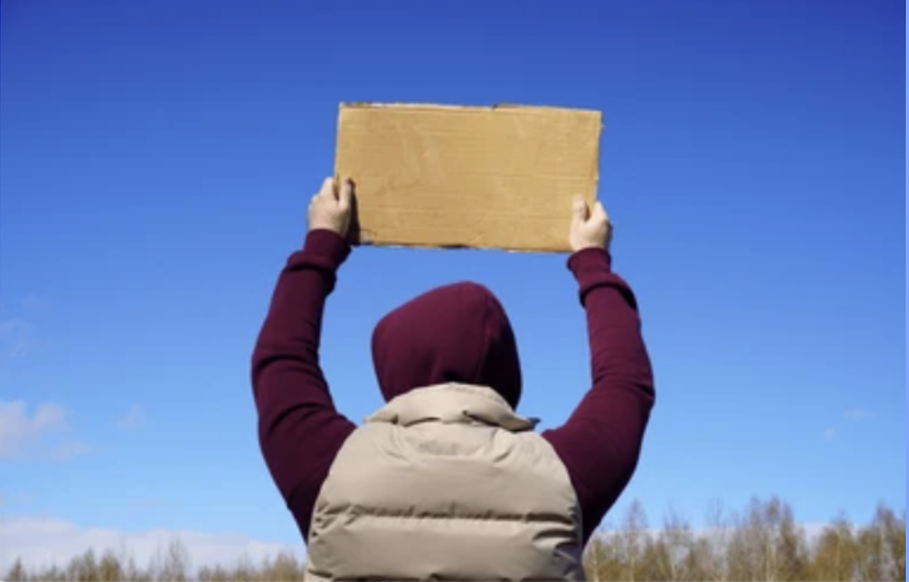 A person in a maroon hoodie and beige vest holds a blank cardboard sign above their head against a clear blue sky, ready to Stand-Up for Democracy.