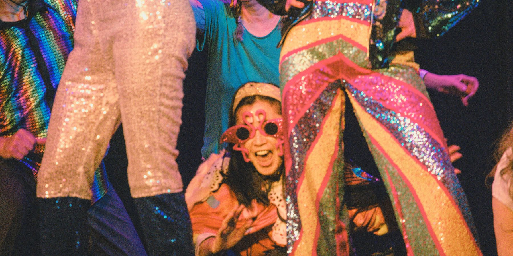People dressed in colorful, sequined costumes pose on stage as the winners of the 2026 KOTO Lip Sync Contest, with one person kneeling in the middle wearing large decorative glasses and smiling.