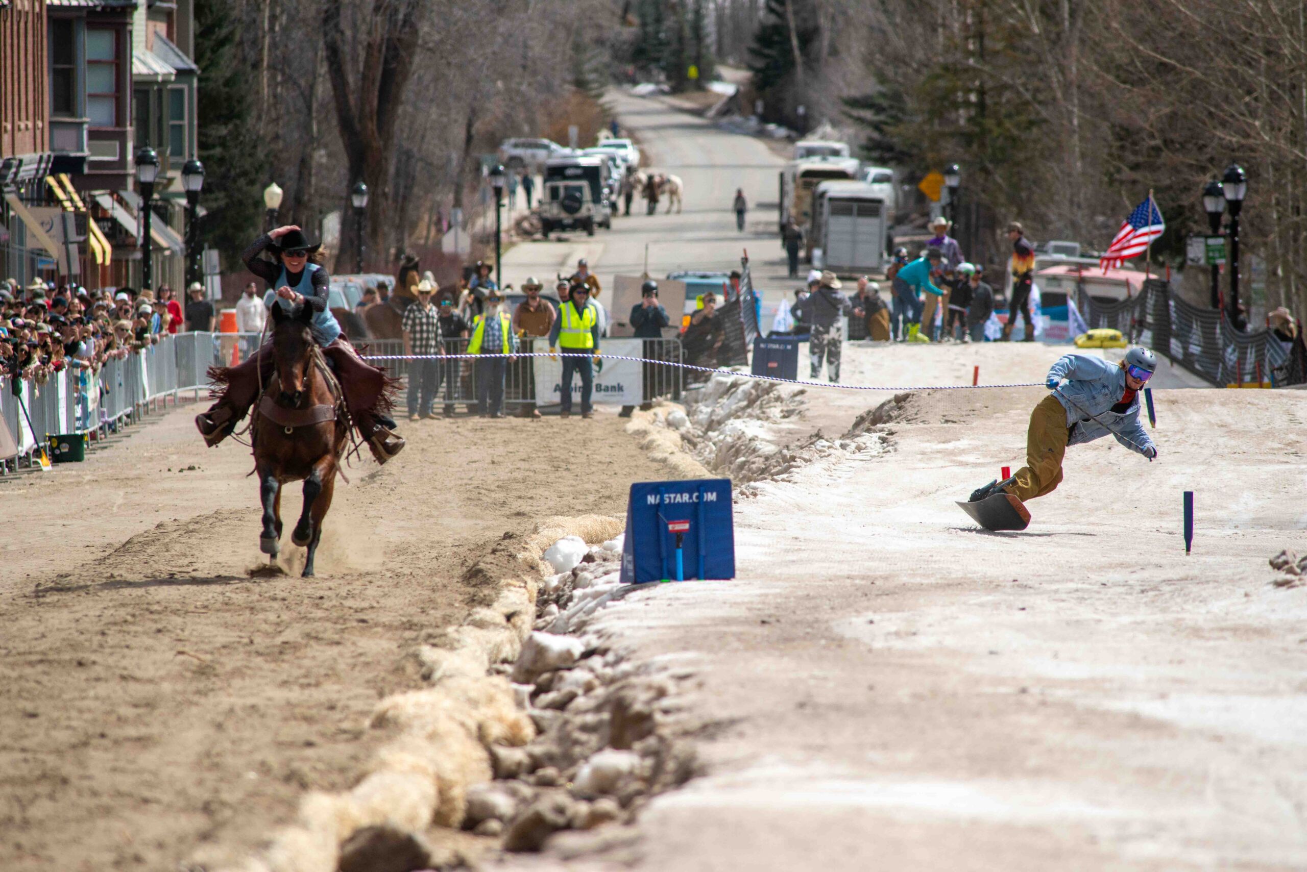 A snowboarder is pulled by a rider on horseback down a snowy track along Main Street during a skijoring event, while spectators watch from behind barriers.