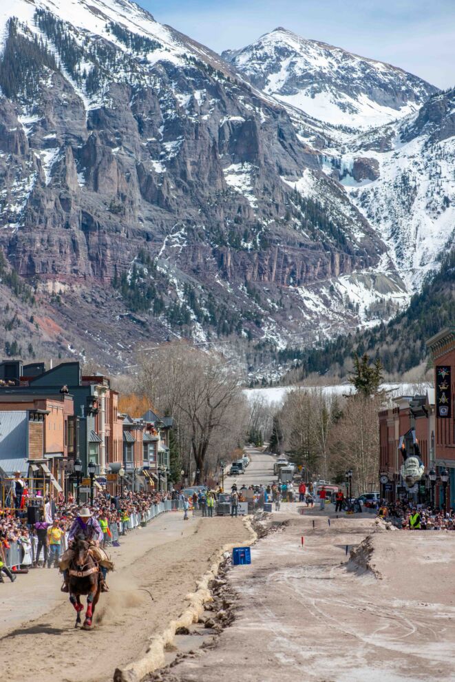 A rider on horseback races down Main Street lined with spectators, with snow-capped mountains and historic buildings in the background, capturing the spirit of skijoring.