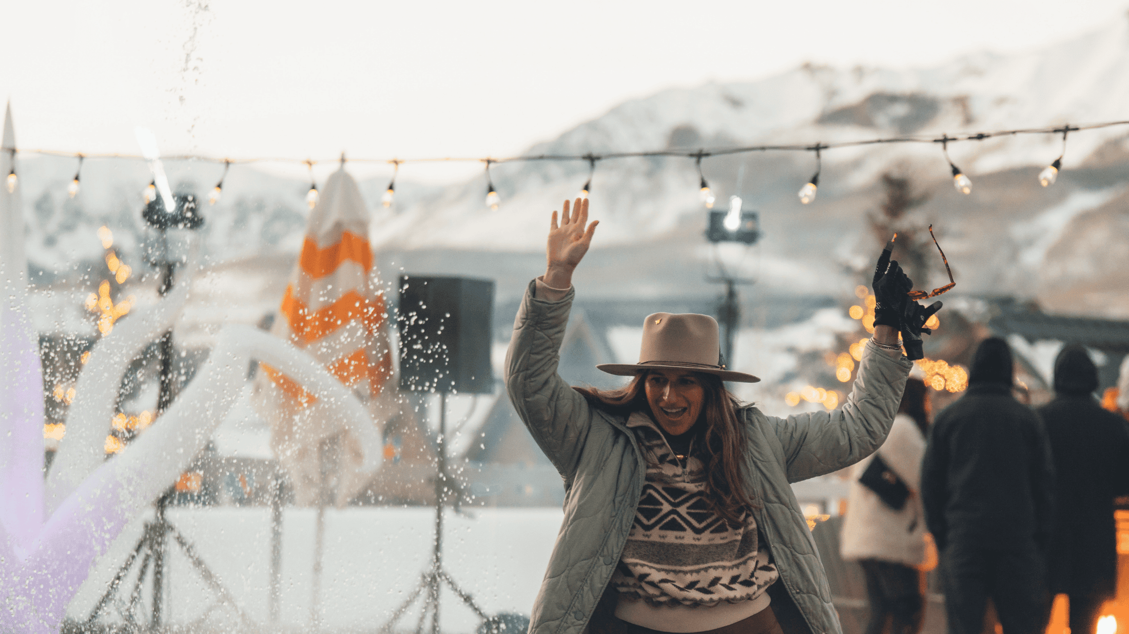 Woman wearing a hat and winter clothes raises her hand and smiles outdoors at Cowboys & Cocktails, with snow-covered mountains, string lights, and other people in the background.