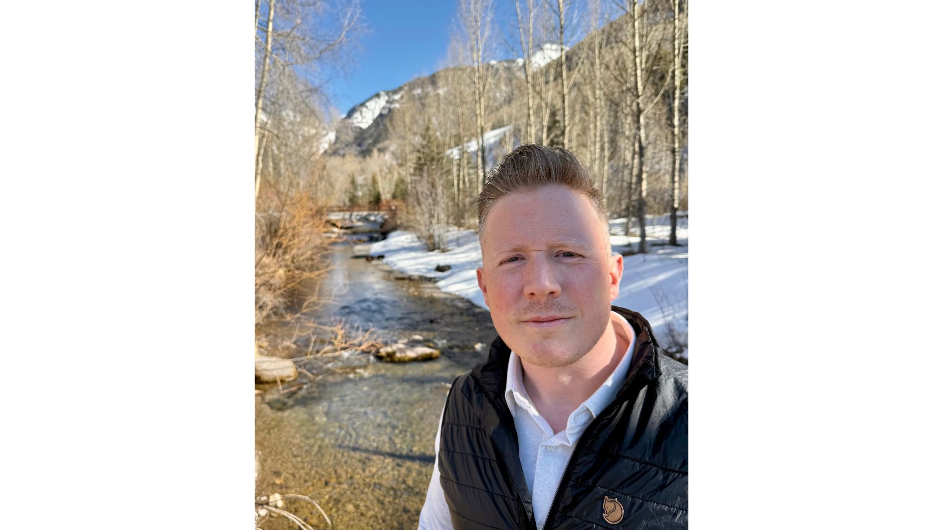 Lane Masters stands by a clear creek with snow on the ground, trees around him, and mountains in the background under a blue sky—a serene moment during the San Miguel County Sheriff election season.