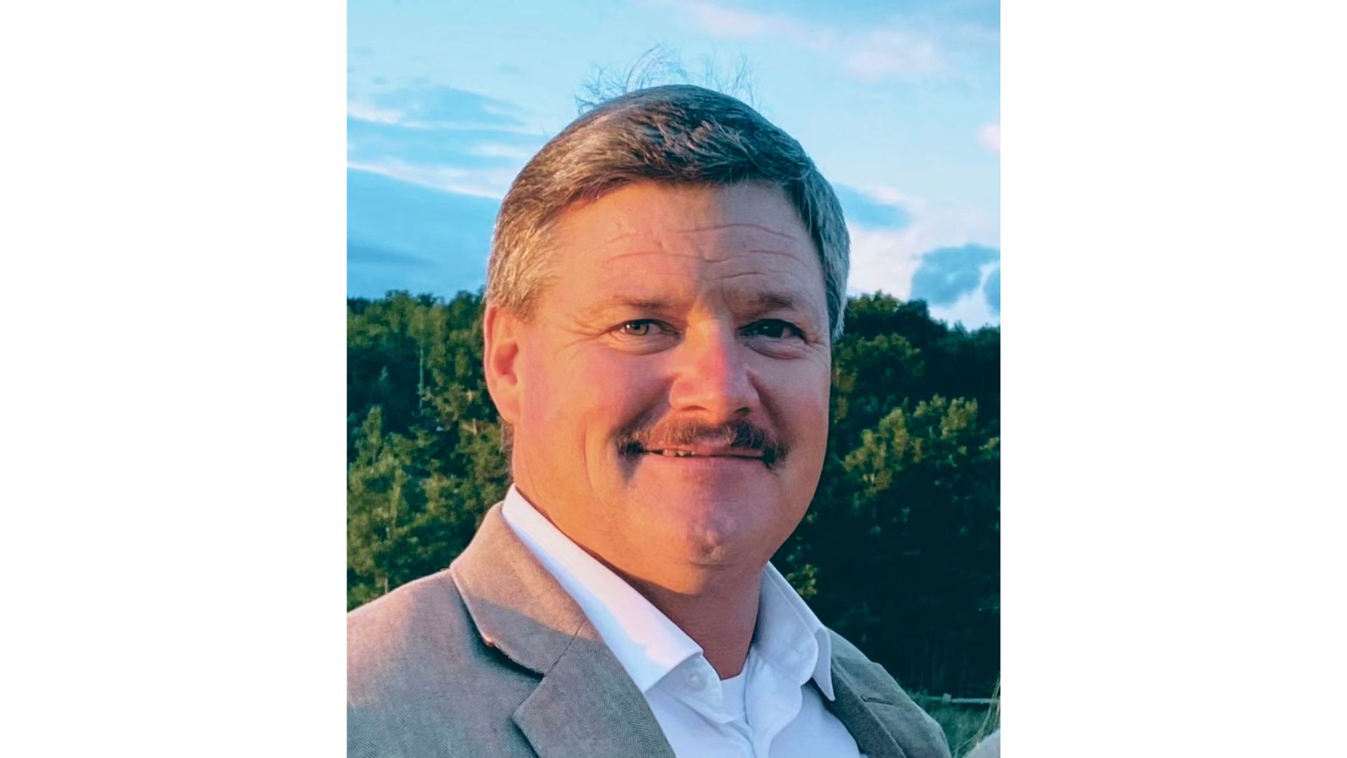 A man with short brown hair and a mustache, wearing a light-colored suit jacket and white shirt, stands outdoors with trees and a blue sky behind him—San Miguel County Sheriff candidate Dan Covualt during the sheriff election campaign.