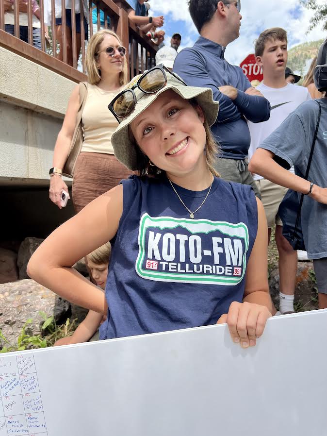 Young woman wearing a bucket hat and KOTO-FM Telluride shirt smiles at the camera, holding a whiteboard; people gather around outdoors at a KOTO News event.