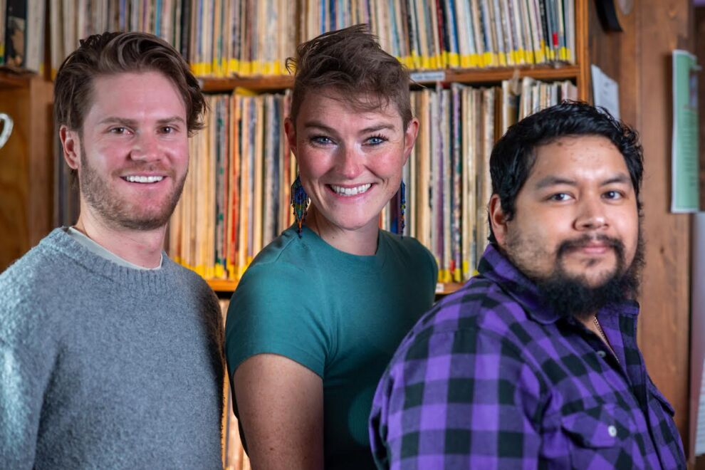 Three people stand in front of shelves filled with records, smiling at the camera during a KOTO News segment.