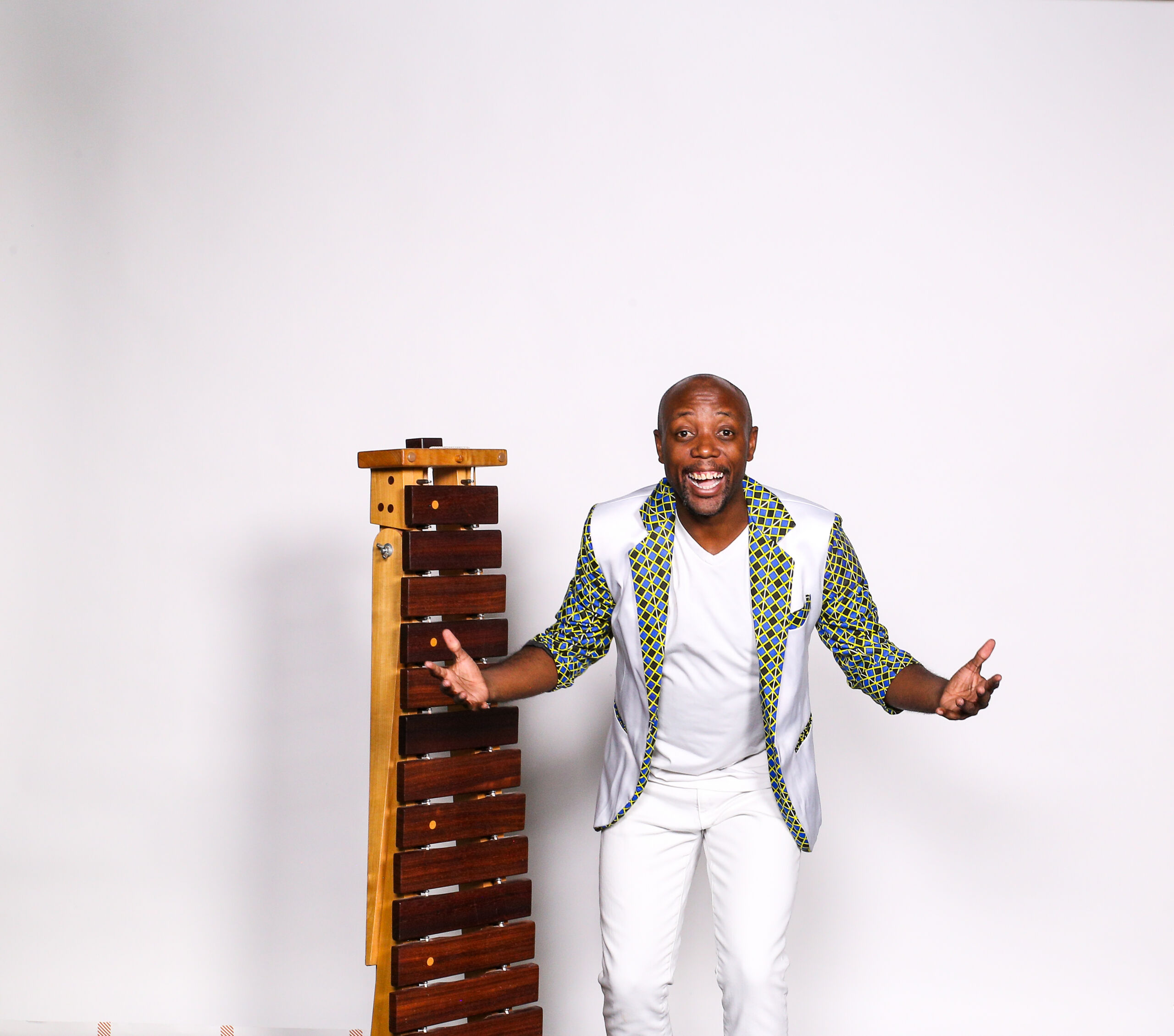Blessing Chimanga, a man in a patterned jacket and white pants, stands beside a wooden xylophone against a plain white background, gesturing with open arms and smiling—capturing the joyful spirit of Zimbabwe's live music.