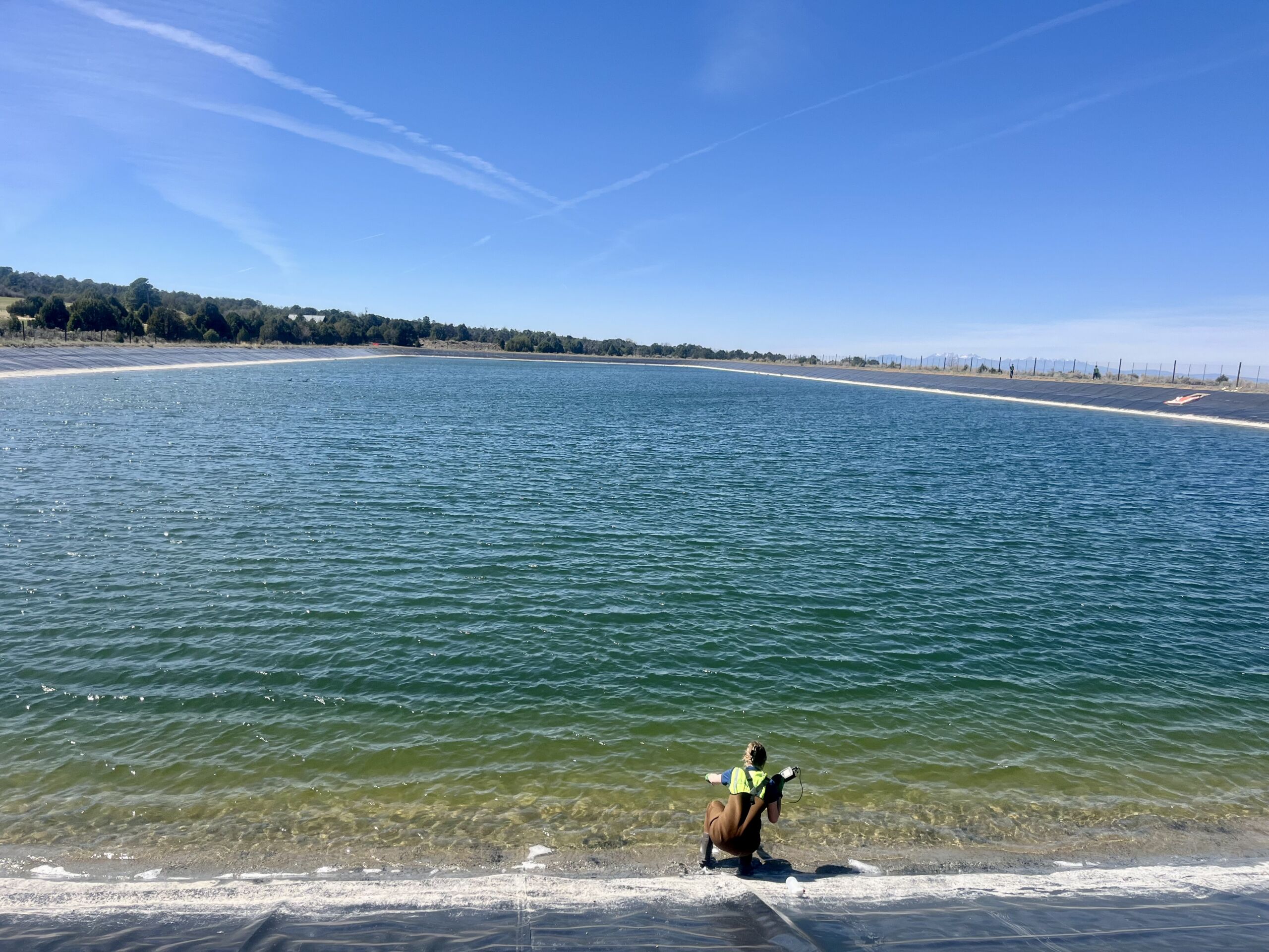 A person in a safety vest crouches by the edge of Reservoir Number 2, observing the calm waters and innovative floating solar panels beneath a clear blue sky.
