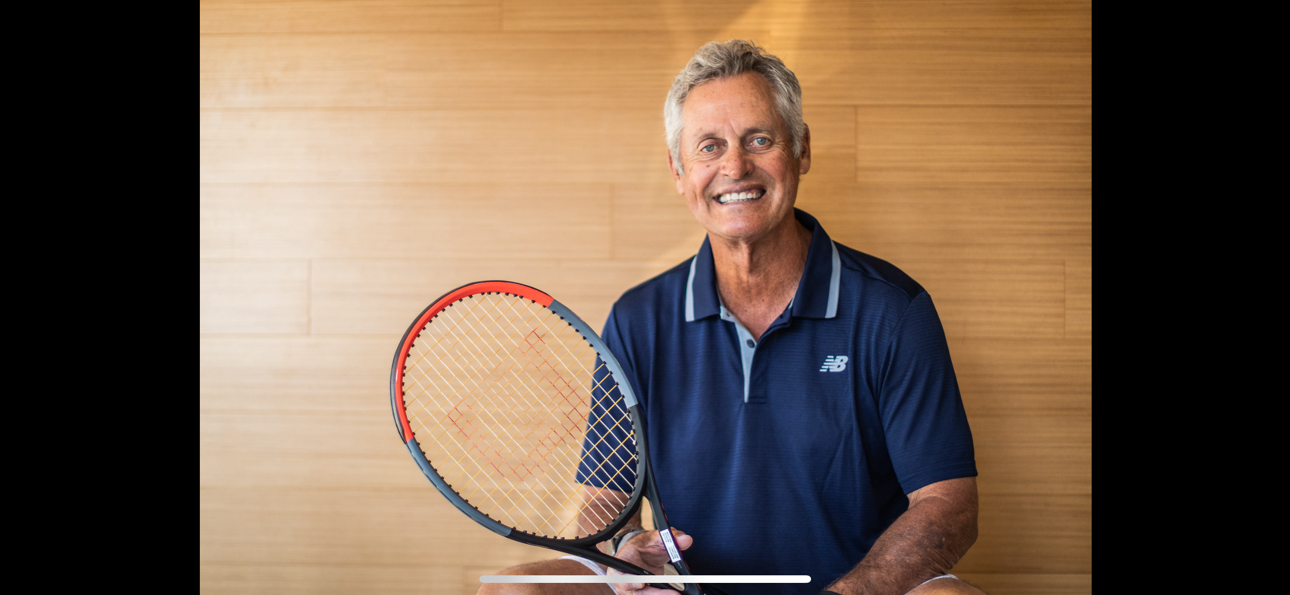 Smiling older man in a navy shirt holding a tennis racket, sitting in front of a light wood background—perfectly capturing the spirit of Tennis Coaching with Tom Gullikson.