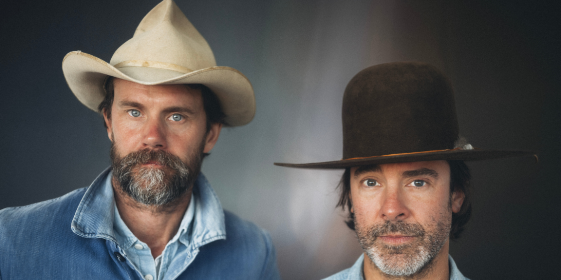 Walker Young and Ben Musser, both in wide-brimmed hats and denim shirts, pose in front of a neutral background, capturing the spirit of Music on the Green.