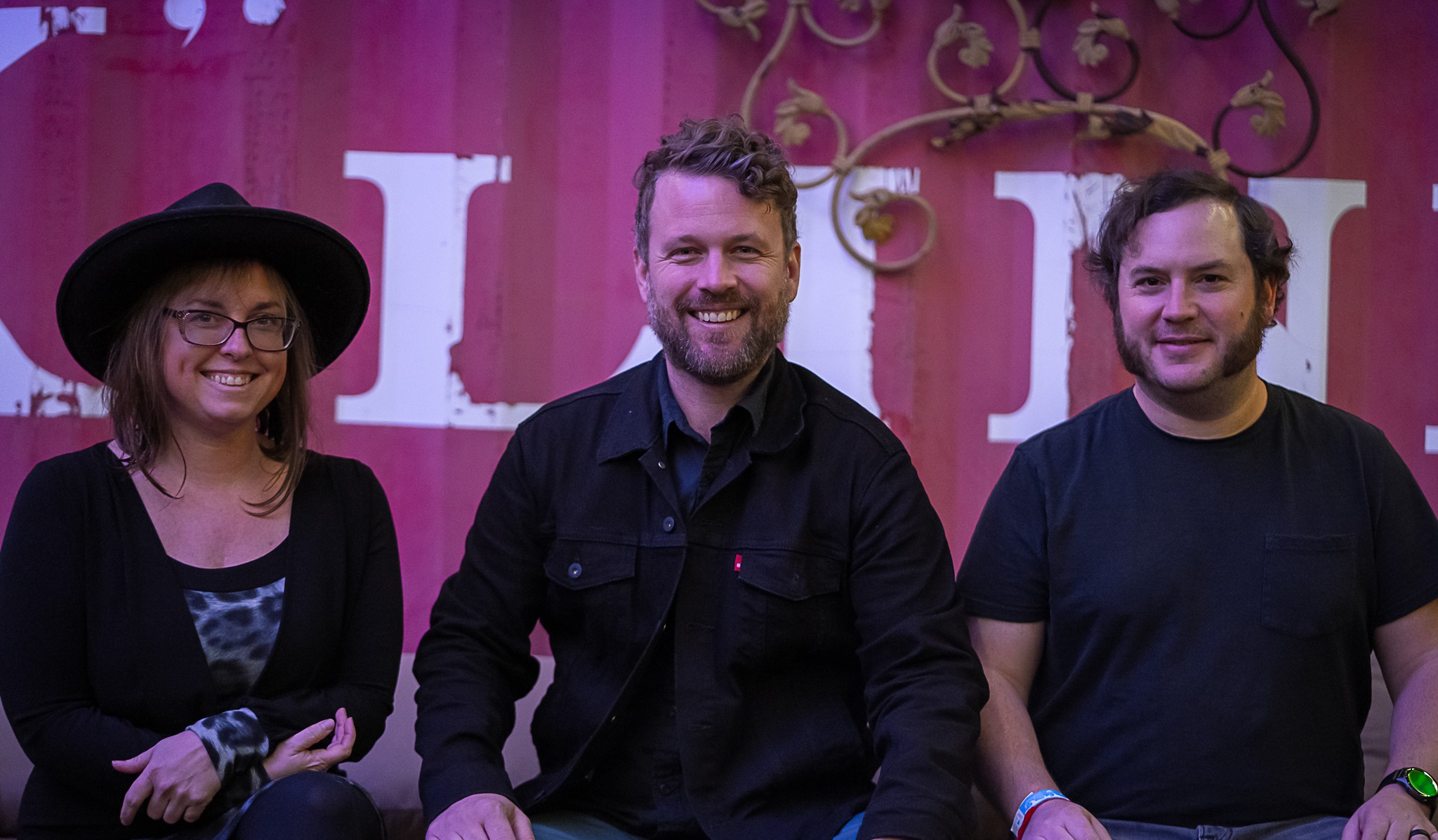 Three people sit side by side in front of a pink and white wall. The woman on the left wears a hat and glasses; the two men on the right, part of Jon Stickley Trio, wear casual dark clothing and smile at the camera after their live music set.