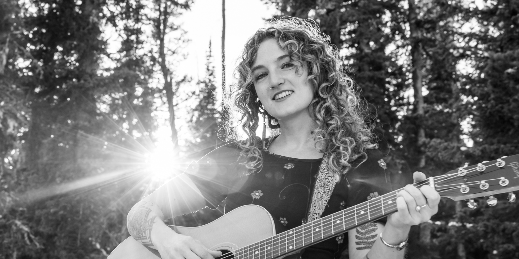 A woman with curly hair plays an acoustic guitar outdoors at Music on the Green, sunlight shining through trees in the background, reminiscent of Madeline Hawthorne's vibrant performances.