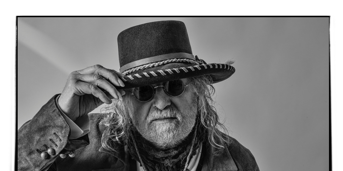 An older man with long hair and a beard, reminiscent of Ray Wylie Hubbard, wears sunglasses and a wide-brimmed hat, tilting the hat with one hand. He has a serious expression and is dressed in layered clothing.