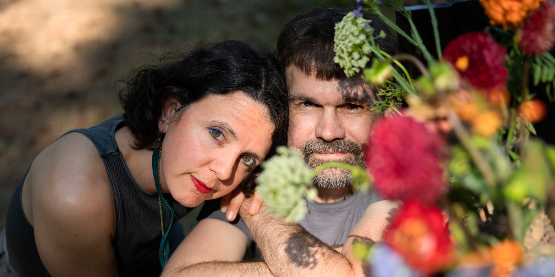 A woman and a man sit closely together outdoors, partially obscured by colorful flowers in the foreground, enjoying the vibrant atmosphere of Music on the Green with Sway Wild performing nearby.