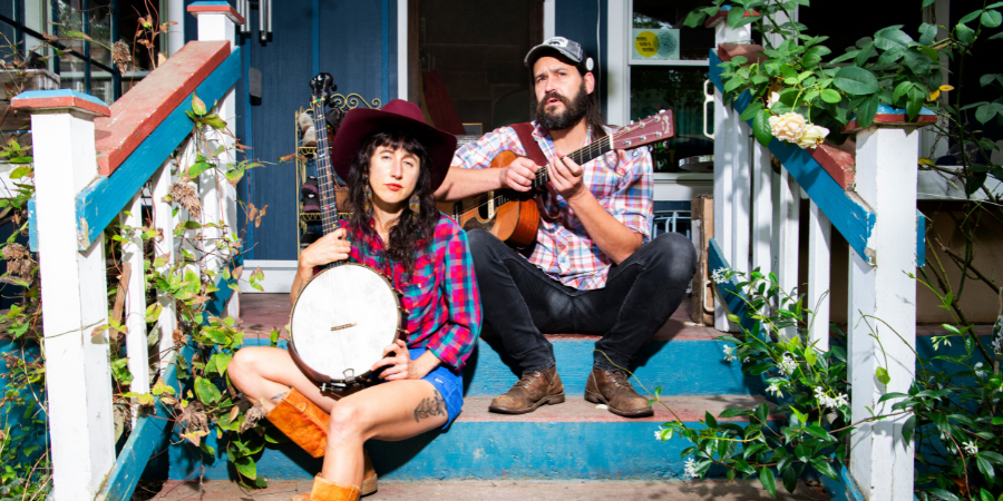 Two people sit on outdoor steps; one holds a banjo, the other plays a guitar. Both wear plaid shirts and hats, as greenery surrounds the steps—capturing the essence of live music with The Lowest Pair during Music on the Green.