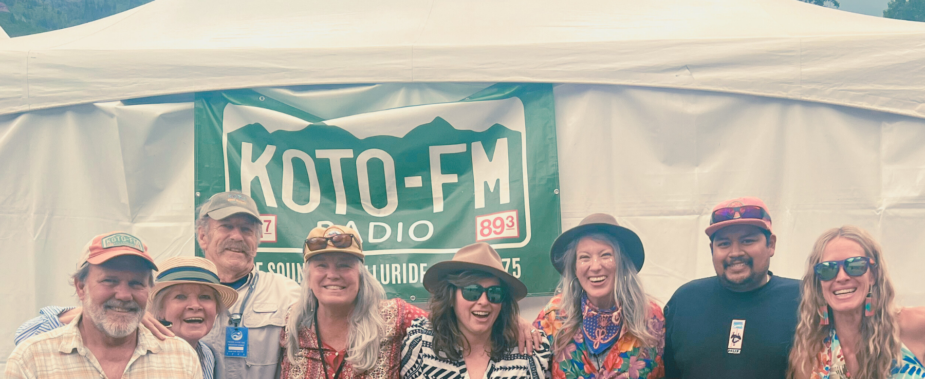 A group of nine people stand and smile in front of a white tent with a green KOTO-FM radio banner displayed behind them.