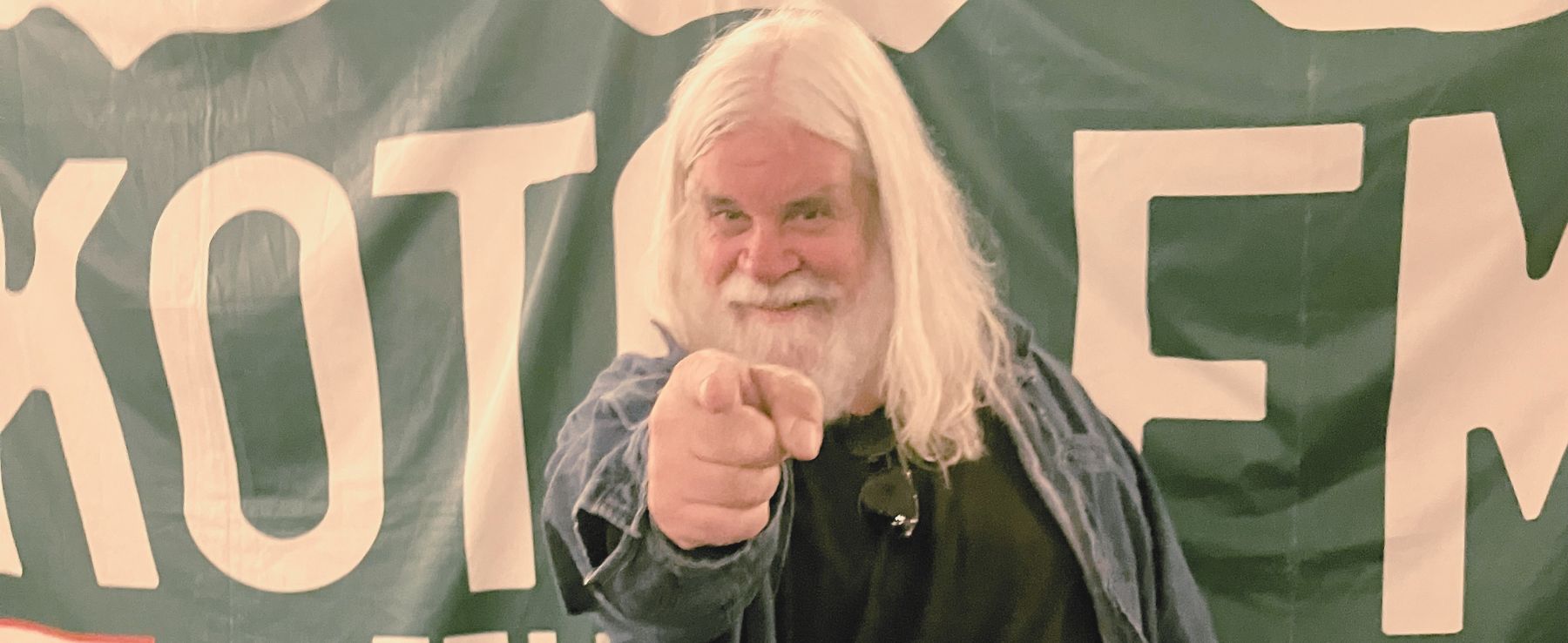 An older man with long white hair and a beard points toward the camera, standing in front of a banner with large, partially visible letters.