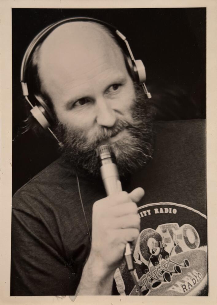 Black and white photo of a bearded man wearing headphones, holding a microphone, and wearing a BF Deal t-shirt with a radio station design.