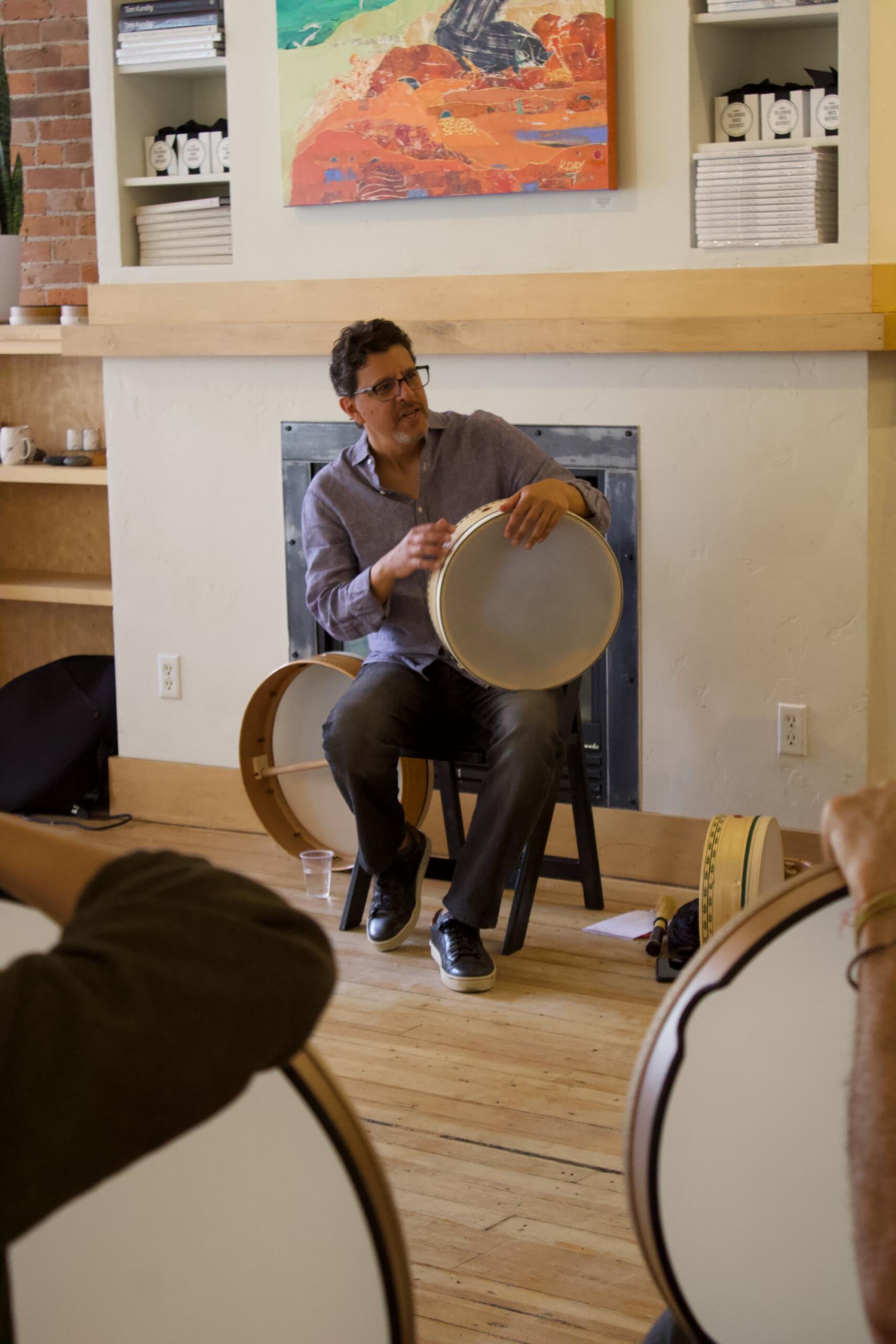 A man sits on a chair playing a frame drum during a Frame Drum Workshop, with two others holding similar drums in the foreground in a room with wooden floors and shelves.