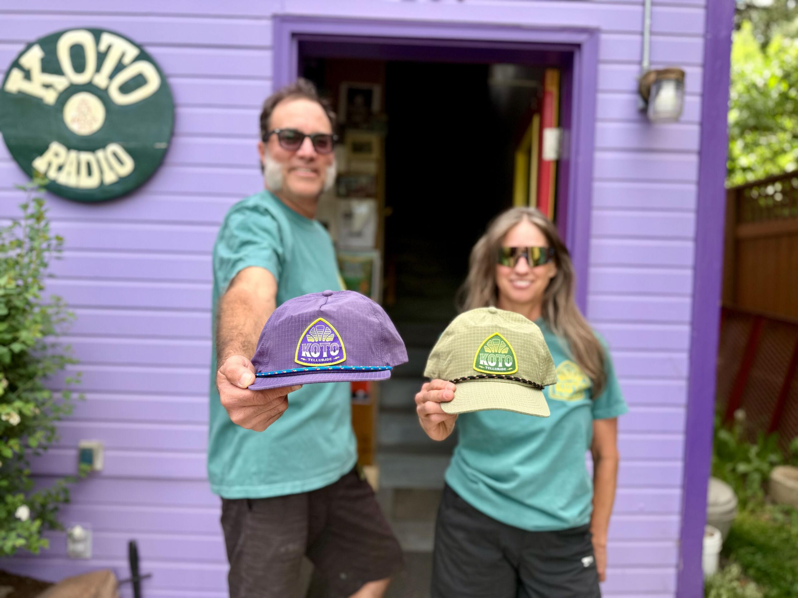 Two people in teal shirts stand outside a purple building labeled KOTO RADIO, each holding a KOTO Radio hat toward the camera.
