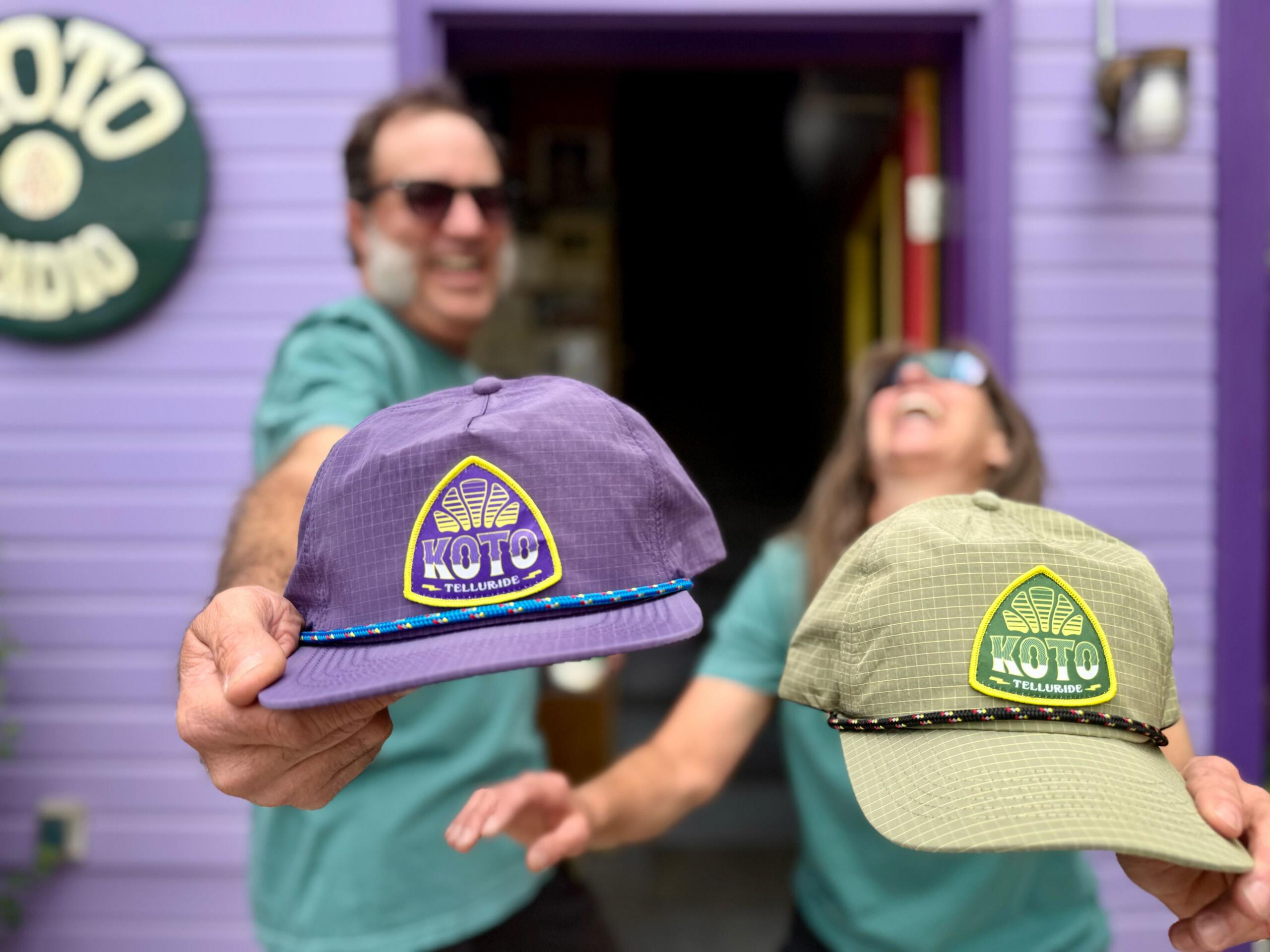 Two people hold out KOTO Telluride hats toward the camera, smiling and standing in front of a purple building with a KOTO studio sign.