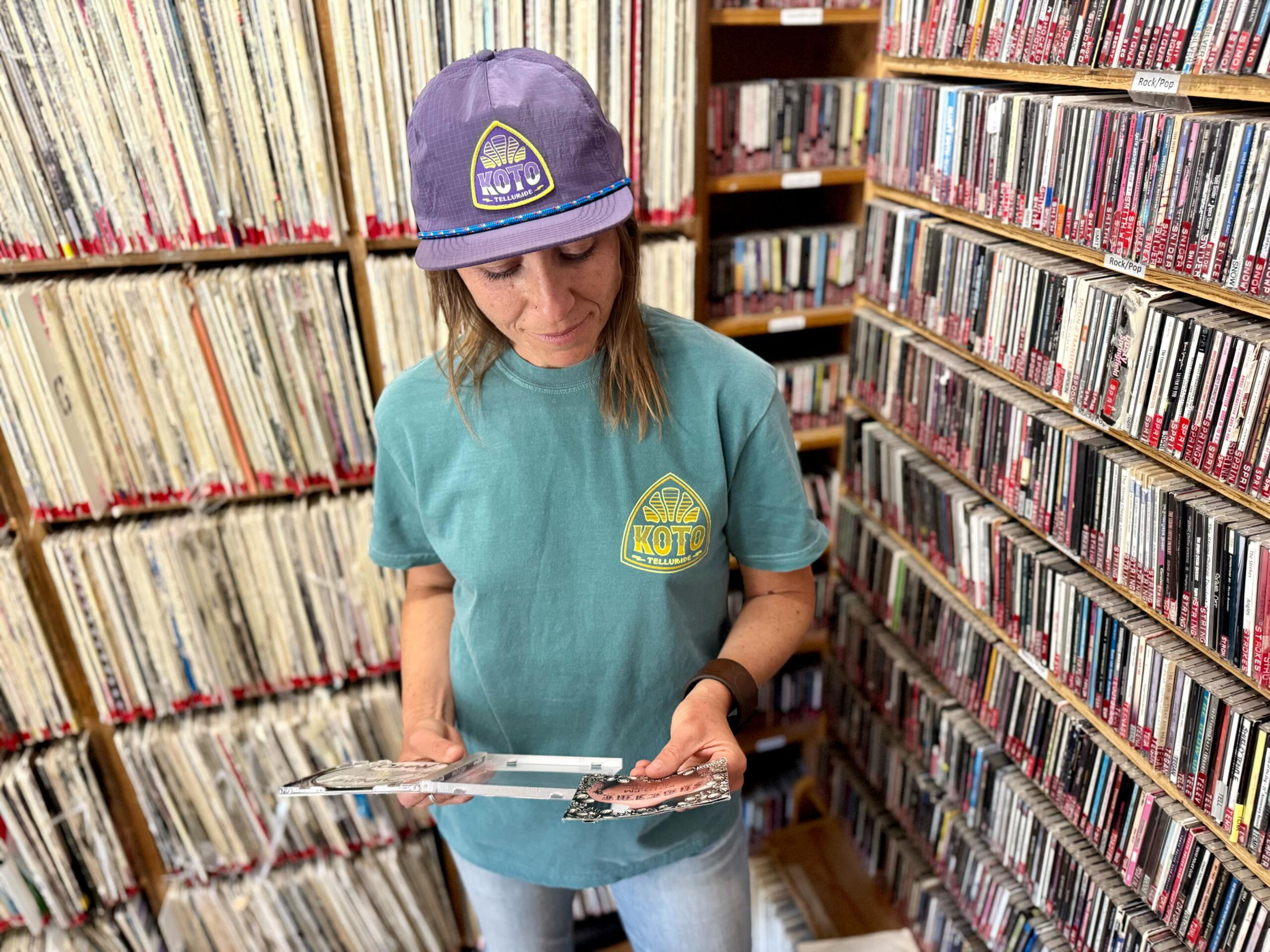 A person wearing a purple hat and teal shirt stands in a room filled with shelves of vinyl records and CDs, looking at album covers in their hands.