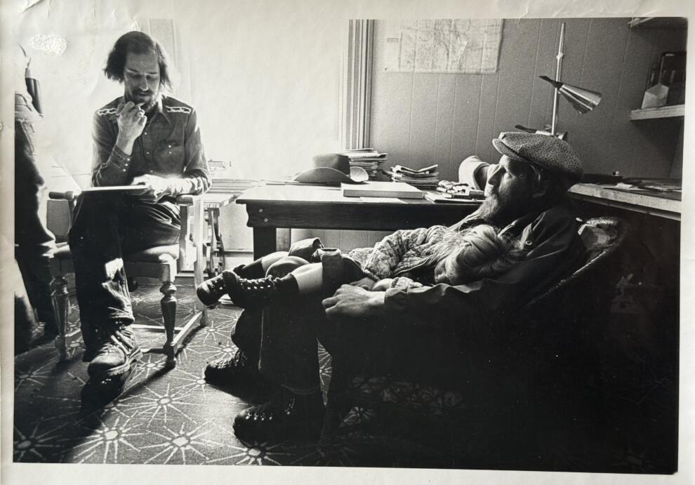 Two men sit in an office; one reads a document, while the other relaxes in an armchair with a child asleep on his lap. Desks, papers, and a lamp hint at work discussions—perhaps even a recent BF Deal—in the background.