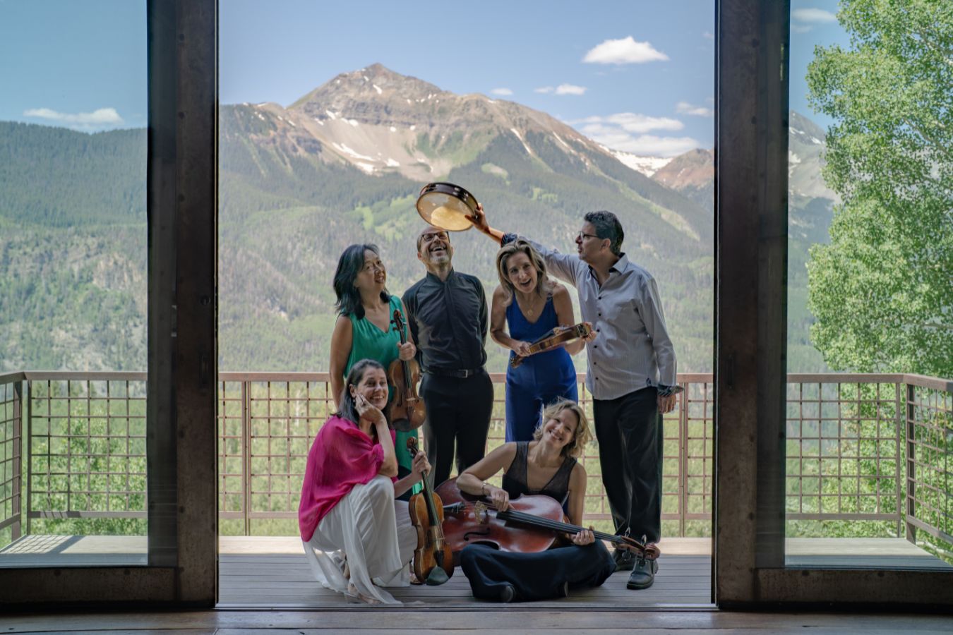 Six musicians pose on a balcony, their string instruments and tambourine ready, as if they've just stepped out of a Music Room, with a scenic mountain landscape unfolding in the background.