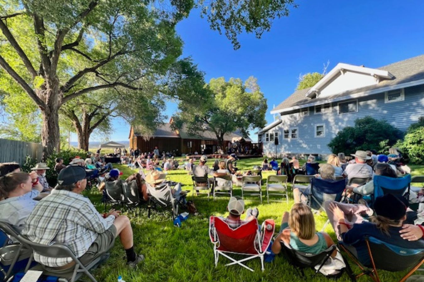People sit on lawn chairs in a shaded backyard during a sunny day, enjoying relaxing music as they face a small stage where a gathering or event appears to be taking place.