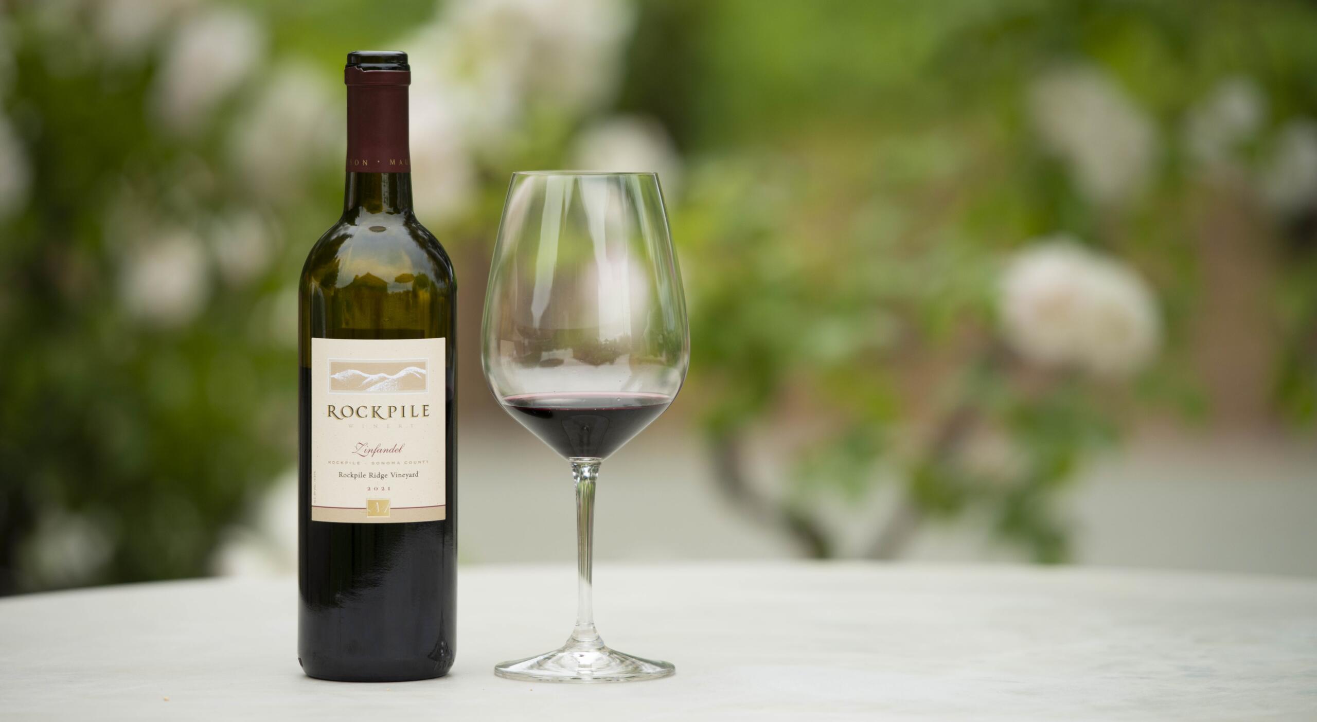 A bottle of Rockpile Zinfandel by Clay Mauritson and a glass of red wine sit on a white table, with a blurred green outdoor background—perfect for your next Winemaker Hike or to meet the winemaker in style.