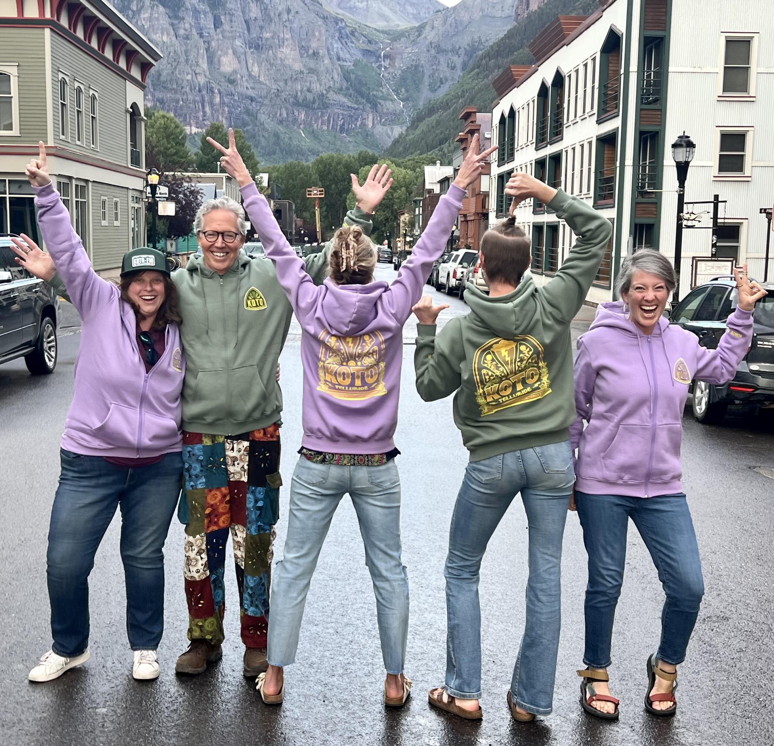 Five people wearing colorful hoodies and jeans pose energetically on a street in a mountainous town, celebrating Bootsie's Bops’ new music. The buildings and scenic mountains in the background capture the lively June 2026 vibe.