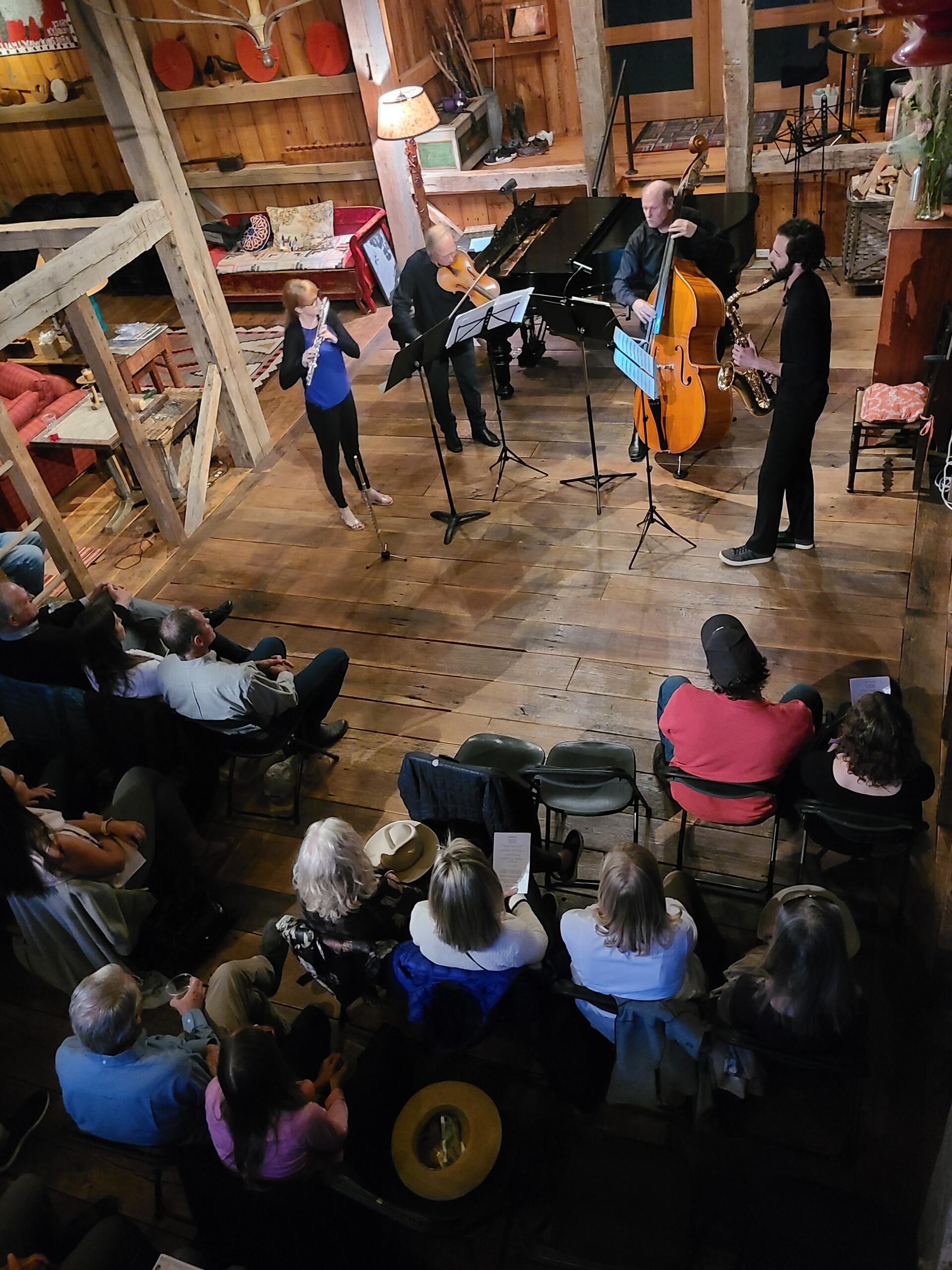 A small ensemble of four musicians performs on a wooden stage in front of an audience during Local Artists Night at a rustic indoor venue.