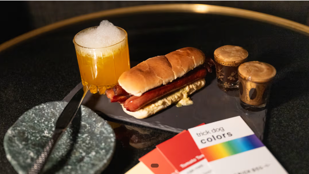 A hot dog in a bun, two small espresso drinks, a yellow cocktail with foam, and color swatches on a black table set the scene for the vibrant Trick Dog Takeover at Timber Room—complete with a marble plate and knife.