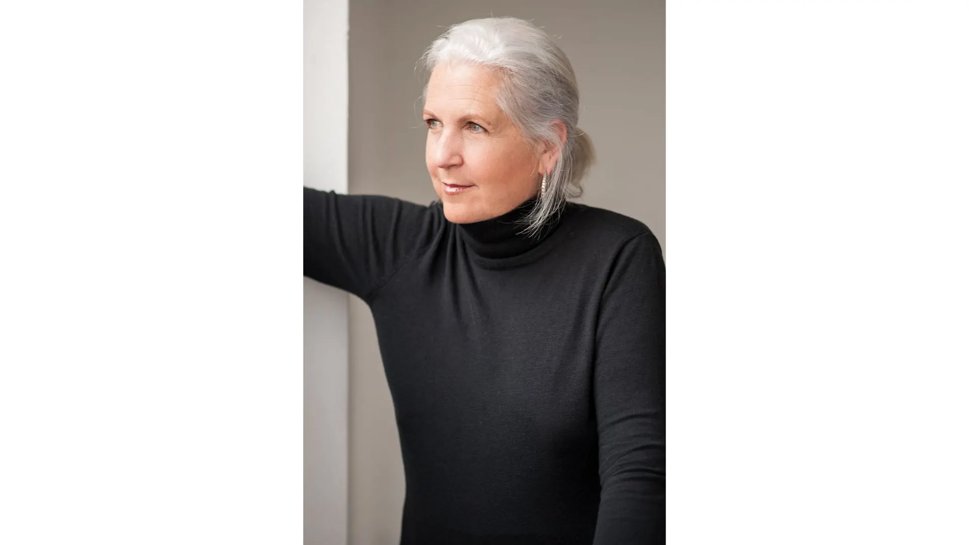 Woman with gray hair wearing a black turtleneck sweater stands indoors, looking to the side with a neutral expression—a quiet grace reminiscent of a Terry Tempest Williams character or one of the thoughtful Glorians.
